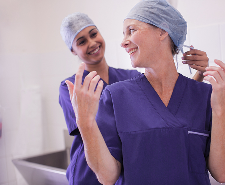 A imagem mostra uma médica auxiliando outra médica com sua roupa em um centro cirúrgico.